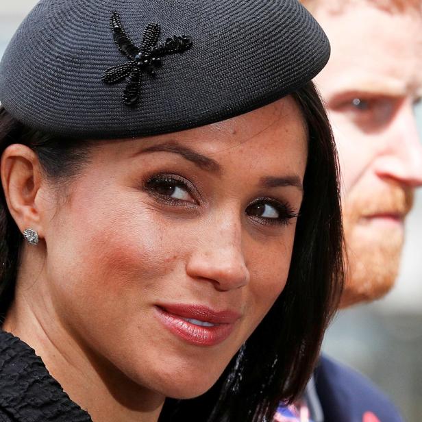 Meghan, die Herzogin von Sussex, mit einem schwarzen Hut und Prinz Harry im Hintergrund.