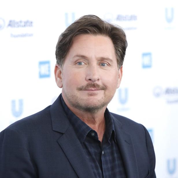 Emilio Estevez posiert vor einem Hintergrund mit Logos.