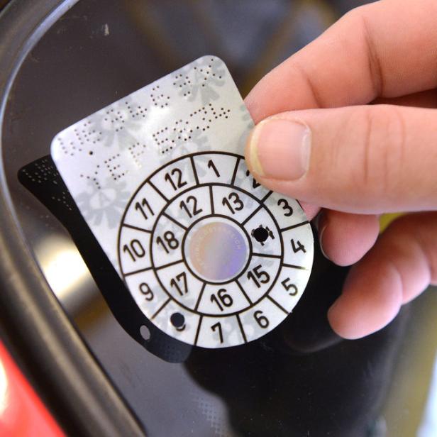 Eine Hand klebt eine österreichische Prüfplakette an eine Windschutzscheibe.