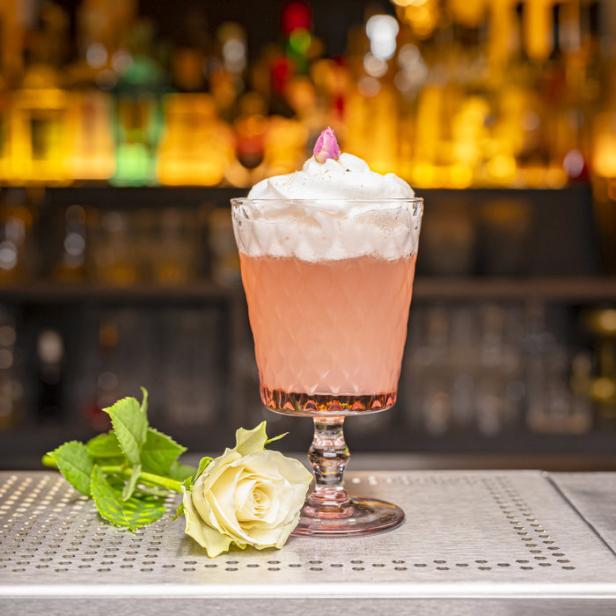 Ein rosa Cocktail mit Schlagsahne und einer Rose steht auf einer Bar.