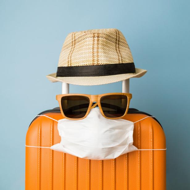 Ein orangefarbener Koffer mit Sonnenbrille, Hut und einer weißen Gesichtsmaske vor blauem Hintergrund.