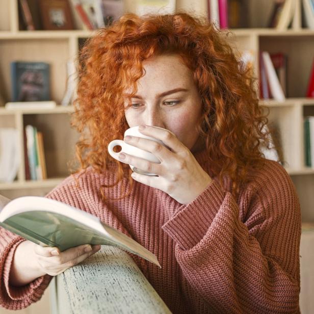 Eine Frau mit roten Haaren liest ein Buch und trinkt eine Tasse Kaffee.