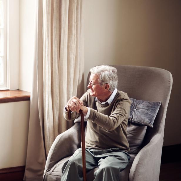 Ein älterer Mann sitzt mit einem Gehstock in einem Sessel und schaut aus dem Fenster.