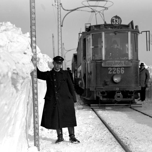 Ein Mann misst die Schneehöhe neben einer Straßenbahn mit der Aufschrift „Franz Josefs Kai 2266“.