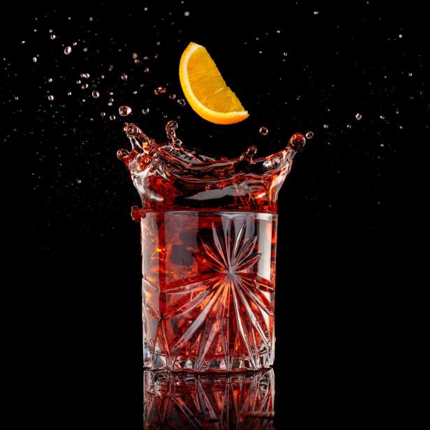 Ein Orangenschnitz fällt in ein Glas mit roter Flüssigkeit und erzeugt eine Fontäne.