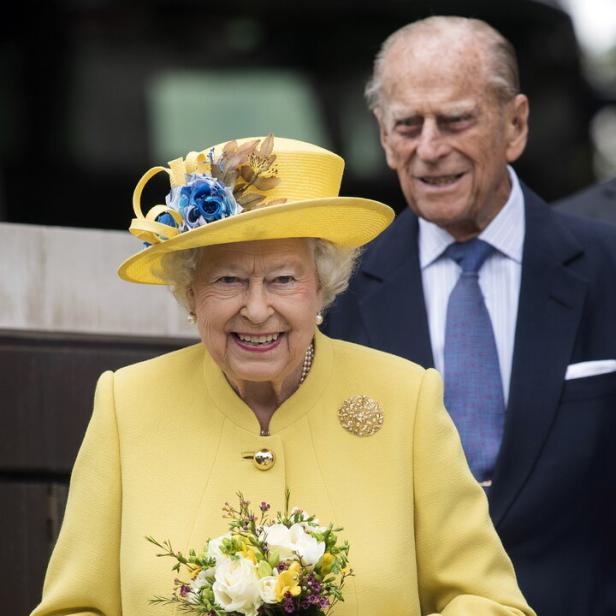 Königin Elisabeth II. lächelt in einem gelben Kostüm mit Blumenstrauß, während Prinz Philip im Hintergrund steht.