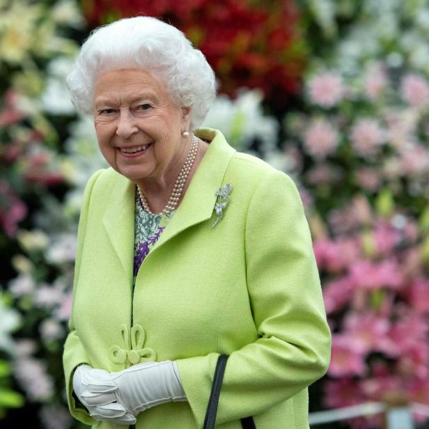 Königin Elizabeth II. lächelt in einem hellgrünen Kostüm mit Brosche vor einem Blumenmeer.