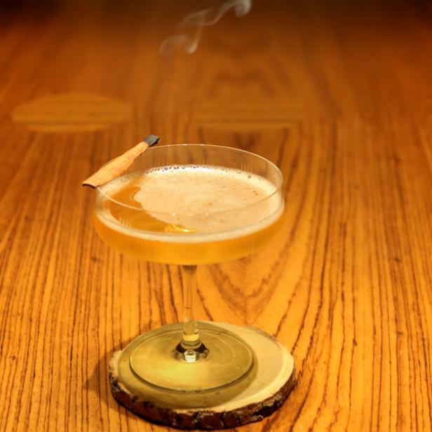 Ein Cocktail mit Zimtstange steht auf einem Holztisch, Rauch steigt auf.