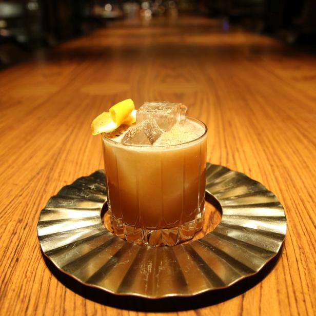 Ein Cocktail mit Eiswürfeln und einer Zitronenzeste auf einem goldenen Untersetzer.