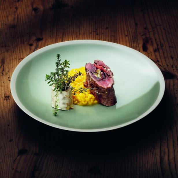 Ein Teller mit Risotto, Fleischroulade und Kräutern auf einem Holztisch.