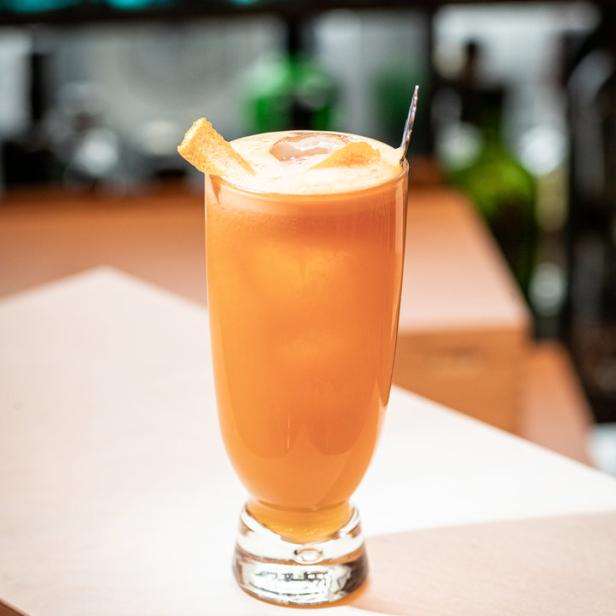 Ein Glas mit einem orangefarbenen Cocktail, garniert mit einer Orangenschale, steht auf einem Tresen.