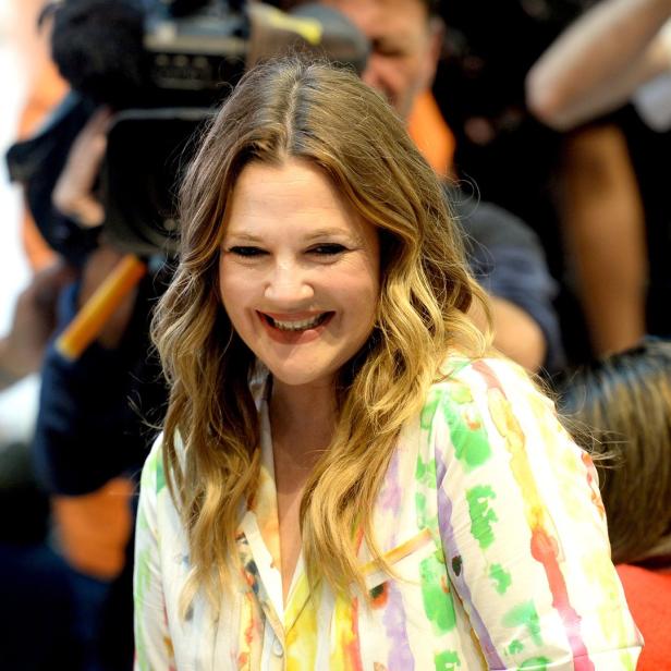 Drew Barrymore lächelt in einem farbenfrohen Hemd umgeben von Kameras.