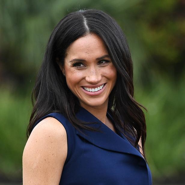 Meghan Markle lächelt in einem ärmellosen, marineblauen Kleid.