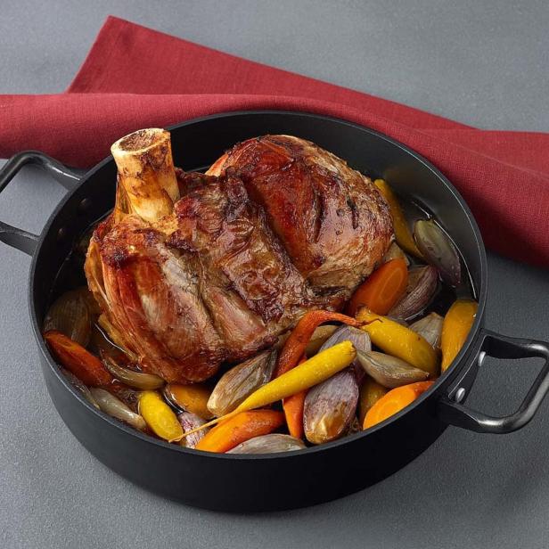 Ein Schweinshaxe mit bunten Karotten und Zwiebeln in einer schwarzen Pfanne.