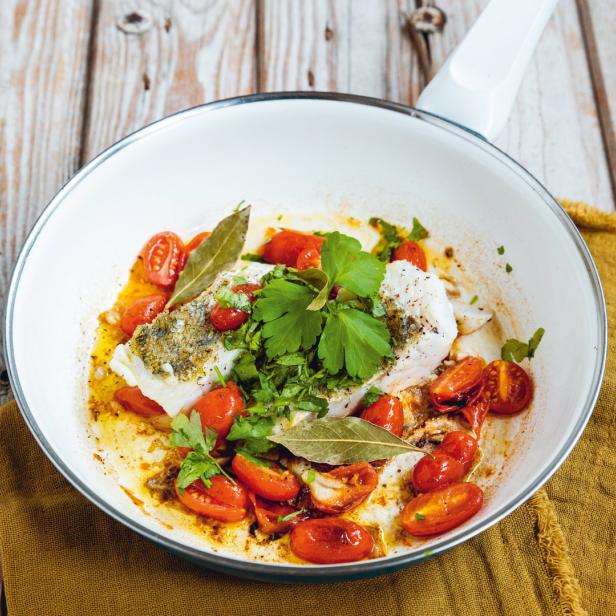 Ein Teller mit Fisch, Kirschtomaten, Kräutern und Lorbeerblättern auf einem Holztisch.
