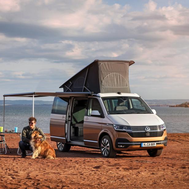 Ein Mann sitzt mit seinem Hund neben einem VW California Campingbus am Strand.