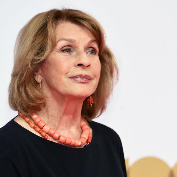 Eine ältere Frau mit blonden Haaren und einer korallenroten Halskette lächelt.