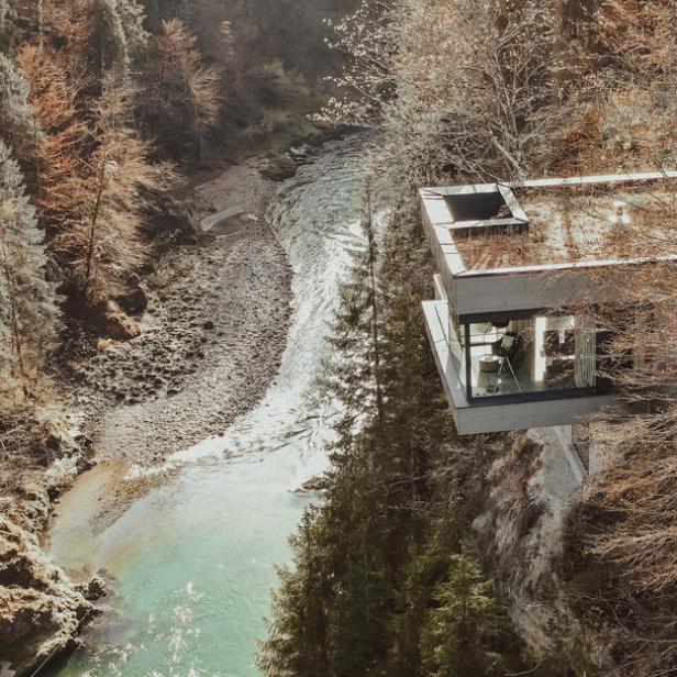 Ein modernes Haus mit Glasfassade hängt über einem Fluss in einer bewaldeten Schlucht.