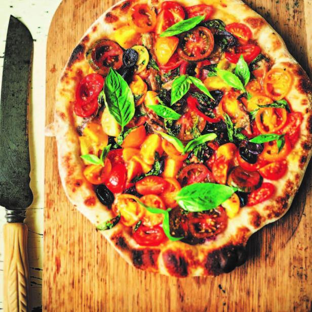 Eine Pizza mit Tomaten, Zucchini und Basilikum auf einem Holzbrett.