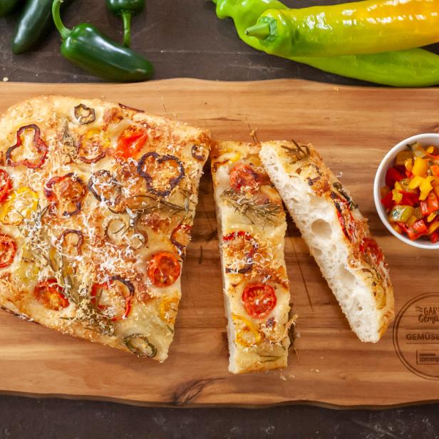 Ein Stück Focaccia mit Gemüse und Kräutern auf einem Holzbrett, umgeben von frischen Paprika.