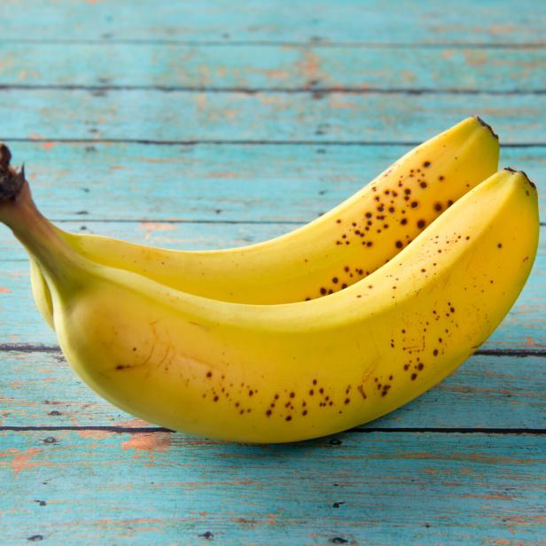 Zwei gelbe Bananen mit braunen Flecken liegen auf einem blaugrünen Holztisch.