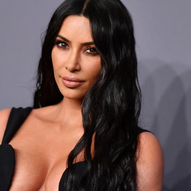 Kim Kardashian posiert in einem schwarzen Kleid vor einem grauen Hintergrund.