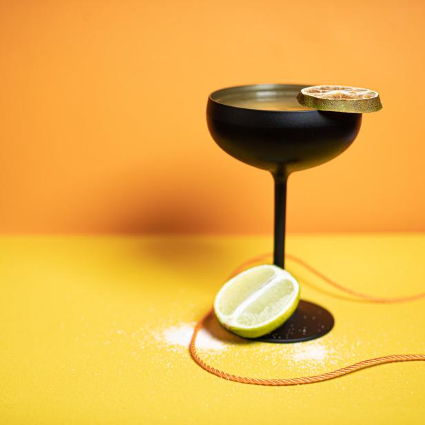 Ein schwarzes Cocktailglas mit Limettenscheibe auf gelbem und orangefarbenem Hintergrund.