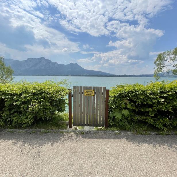 Ein Holztor mit gelbem Schild führt zu einem See mit Bergen im Hintergrund.