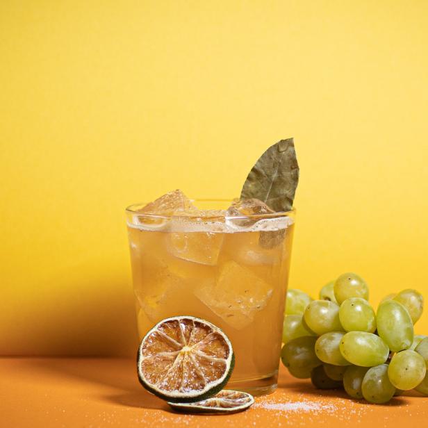 Ein Glas mit einem Getränk, Eiswürfeln, einem Lorbeerblatt und einer Limettenscheibe, daneben eine Traube.