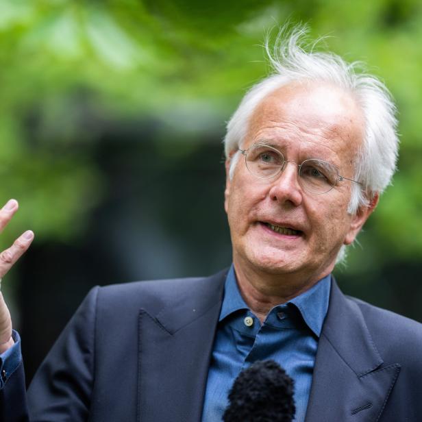 Ein Mann mit weißem Haar und Brille gestikuliert vor einem grünen Hintergrund.