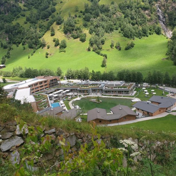 Blick auf ein Hotelresort mit Pool und See in einer grünen Berglandschaft.