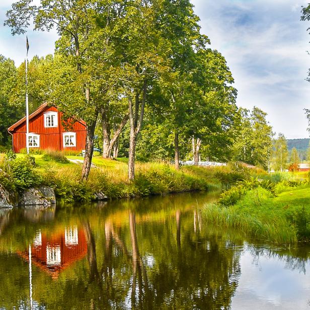 Ein rotes Holzhaus mit weißen Fensterrahmen spiegelt sich in einem ruhigen Fluss.
