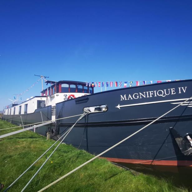 Das Schiff „Magnifique IV“ liegt an einem grünen Ufer vor blauem Himmel.