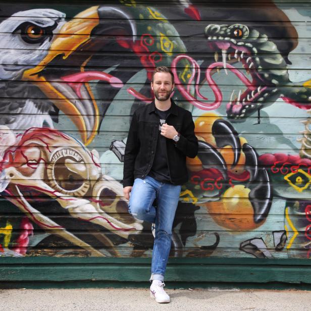 Ein Mann posiert vor einer Wand mit Graffiti von einem Adler und einer Schlange.
