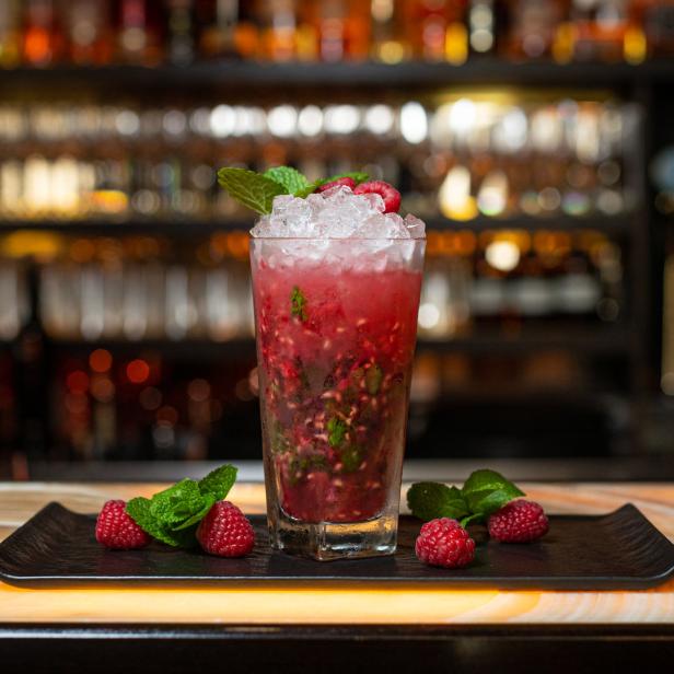 Ein Himbeer-Mojito mit Minze und Eiswürfeln auf einem dunklen Teller an einer Bar.