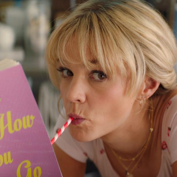 Eine blonde Frau trinkt mit einem Strohhalm und liest das Buch „Careful How You Go“.