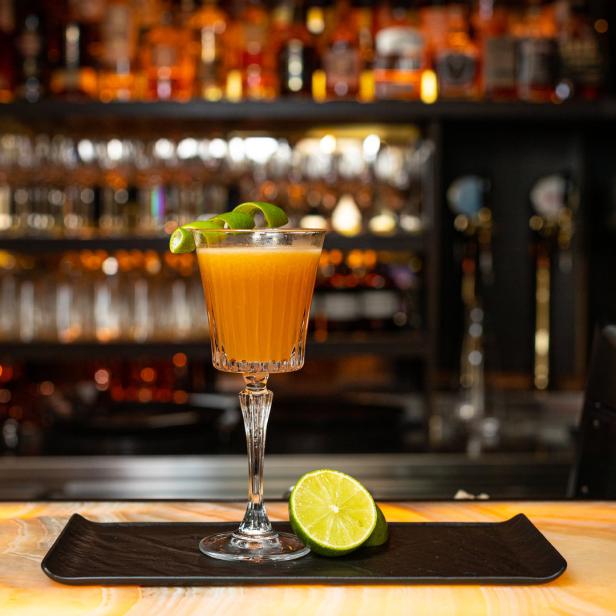 Ein Cocktail mit Limettenspirale steht auf einem Tresen mit einer Limette daneben.