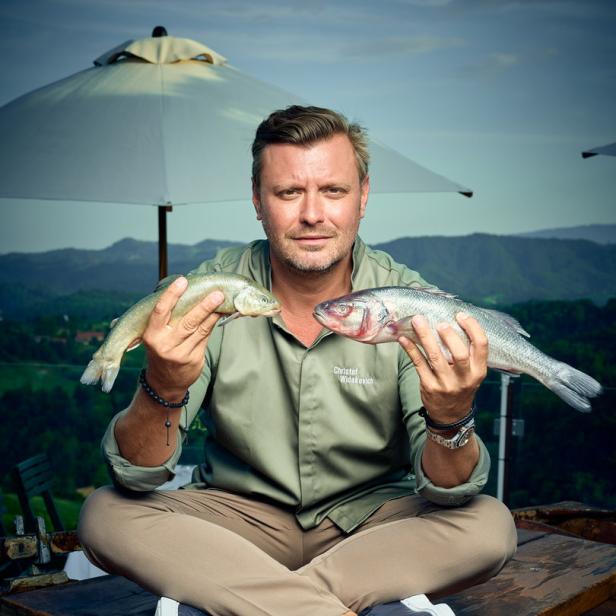 Christof Widakovich sitzt mit zwei Fischen in der Hand vor einer Bergkulisse.