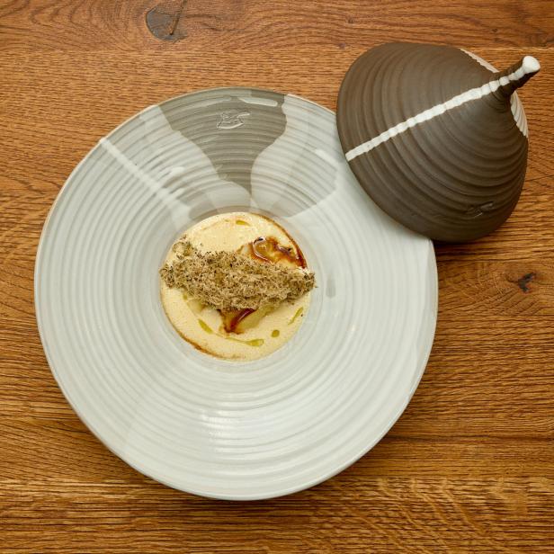 Ein Teller mit einer hellen Suppe, garniert mit Trüffeln und einem dekorativen Deckel daneben.