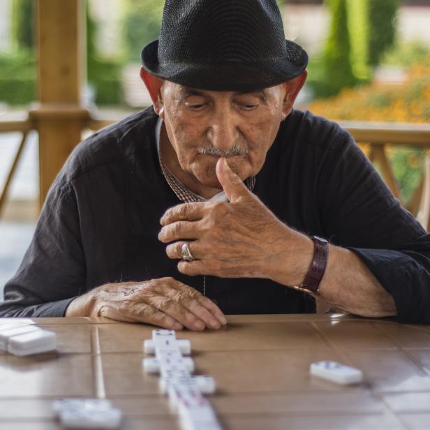 Ein älterer Mann mit Hut spielt Domino auf einer Terrasse.