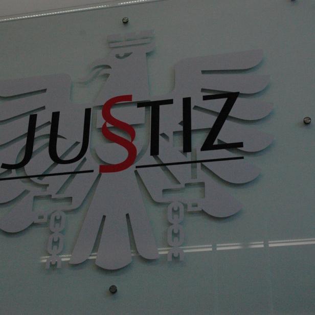 Das Logo der Justiz mit Adler und dem Wort "Justiz", wobei das "S" rot ist, ist an einer Wand befestigt.