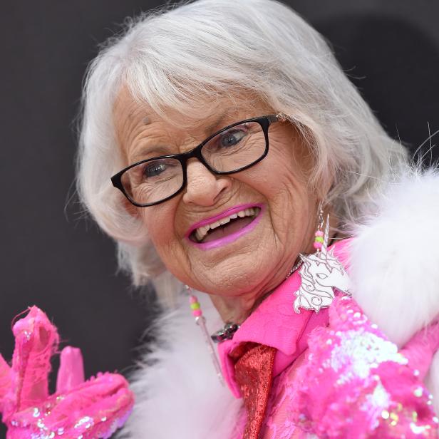Eine ältere Frau mit Brille, pinkfarbenen Accessoires und einem Einhorn-Ohrring lächelt.
