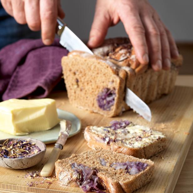 Ein Brot mit roten Zwiebeln wird auf einem Holzbrett angeschnitten.