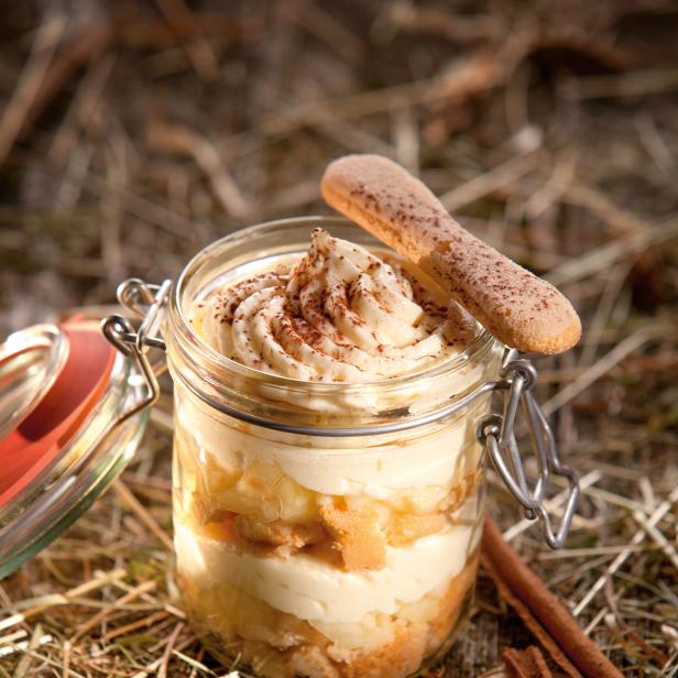 Ein Apfel-Tiramisu im Glas mit Löffelbiskuits und Zimtstangen auf einem Holztisch.