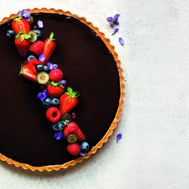 Eine Schokoladentarte ist mit Erdbeeren, Himbeeren, Blaubeeren und lila Blüten verziert.
