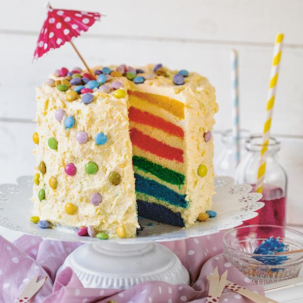 Ein angeschnittener, farbenfroher Regenbogenkuchen mit Zuckerstreuseln und einem kleinen Schirmchen steht auf einem Tisch.