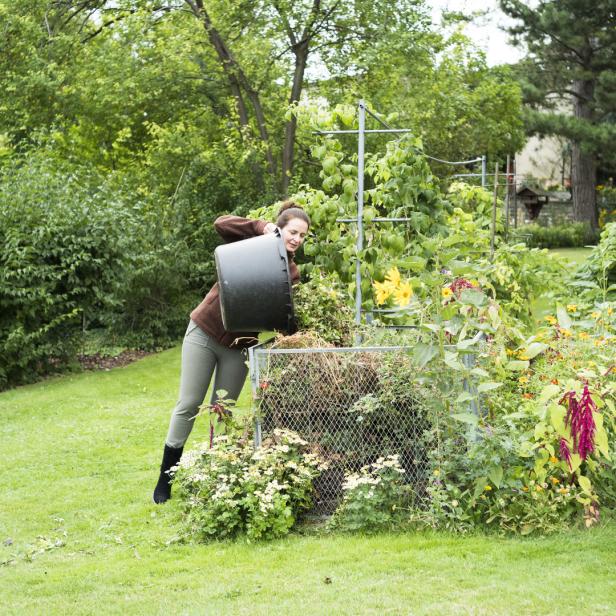 Eine Frau leert einen schwarzen Eimer in einen Komposthaufen im Garten.