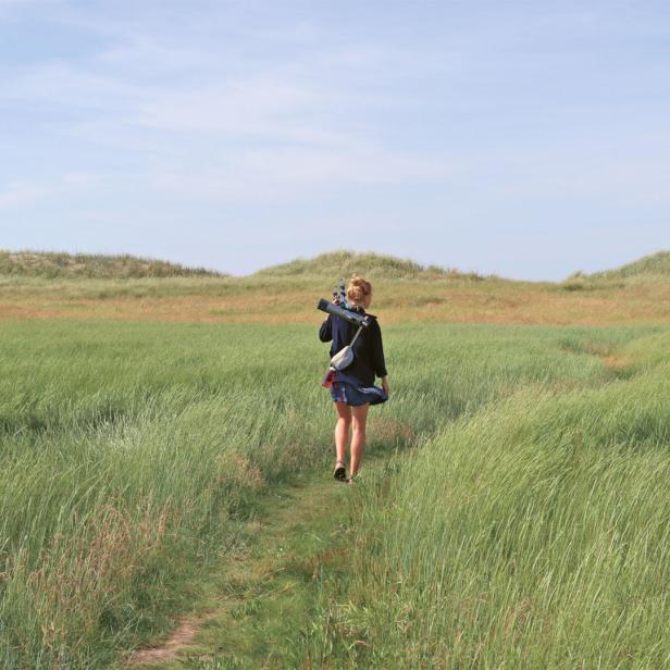 Eine Frau geht mit einem Teleskop über ein Feld in Richtung Dünen.
