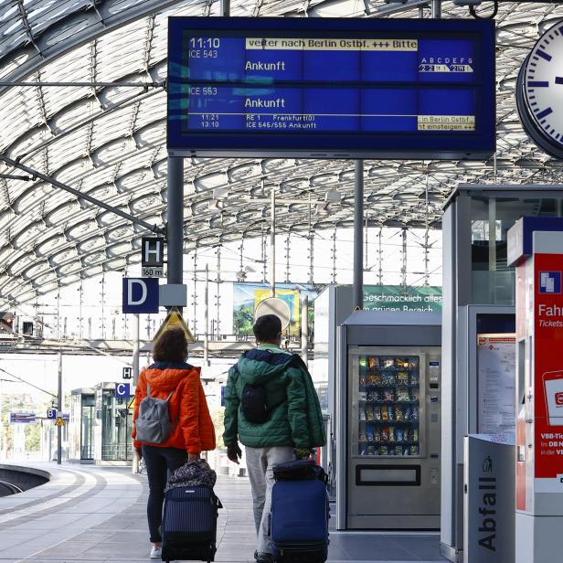 Zwei Reisende mit Gepäck auf einem Bahnsteig mit Anzeigetafel und Fahrkartenautomaten.