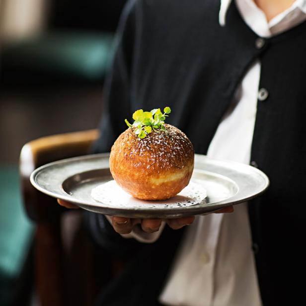 Ein Kellner präsentiert einen mit Puderzucker bestäubten und mit Grünzeug garnierten Krapfen auf einem silbernen Tablett.